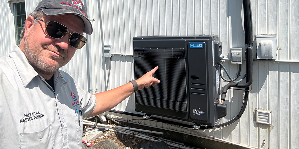 HVAC contractor Mikey Pipes pointing at installed ACiQ heat pump system outside home