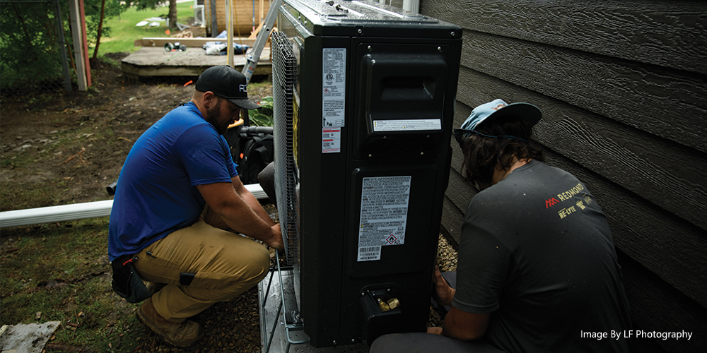 Two HVAC technicians working on an outdoor heating and cooling unit beside a residential home